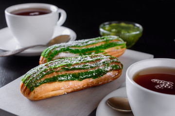 Delicious fresh eclairs garnished on top with stripes of green icing next to a white cups of tea and jam on a black table are ready for eating