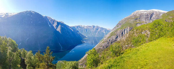 Fototapeta na wymiar An majestic view on Eidfjord from Kjeasen, Norway. Slopes of the mountains are overgrown with lush green grass. Water has dark blue color. Taller parts of the mountains are barren. Sunny and clear day