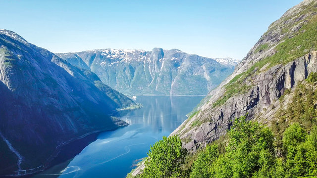 An majestic view on Eidfjord from Kjeasen, Norway. Slopes of the mountains are overgrown with lush green grass. Water has dark blue color. Taller parts of the mountains are barren. Sunny and clear day