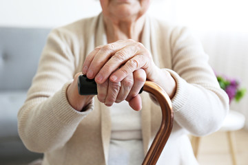 Elderly woman sitting in nursing home room holding walking quad cane with wrinked hand. Old age senior lady wearing beige cardigan, metal aid stick handle bar close up. Interior background, copy space