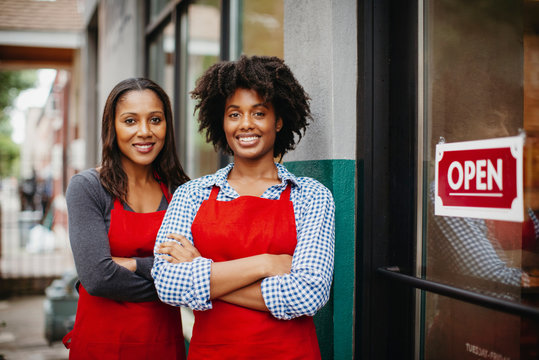 Portrait of smiling female coworkers with arms crossed standing by open sign at cafe