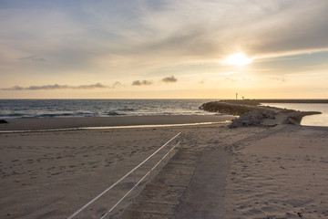 Sunset on empty ocean beach with stone pier. Ocean coast landscape. Evening sky in dusk on seashore. Sun in clouds above the sea. Travel concept. 