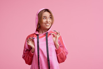 Portrait of a cheerful young girl in pink hoodie clothes looking at camera and winking isolated over pink background