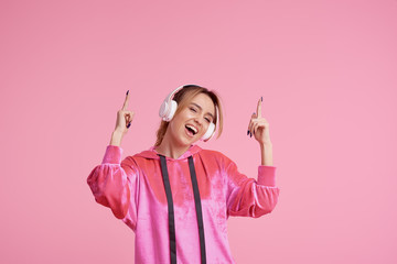 Wireless connection. Portrait of young woman posing isolated over pink background listening fun music with headphones. Shows fingers up. Space for text