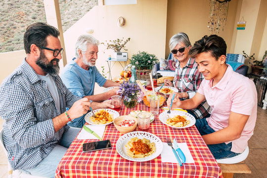 Family people outdoor lunch together on a red table - mixed ages generations enjoy and have fun with italian pasta on the terrace at home - father grandfather and son