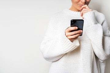 A young woman in a white sweater holds a smartphone in her hands, chooses something looking at the...