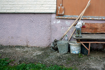 Two old buckets and shovels stand near a bench by the wall of the house. Spring work in the village. Black earth on a spade and green grass in a village in spring.