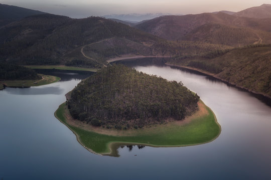Aerial view of wooded island on smooth surface of lake in deserted undulating terrain