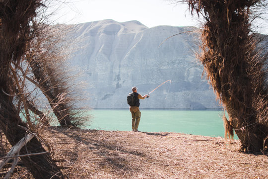 Back view of male with backpack standing on lake shore and fishing on sunny day in nature