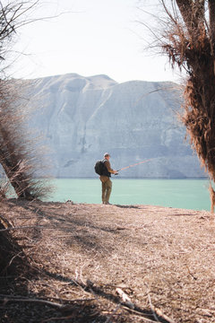 Back view of male with backpack standing on lake shore and fishing on sunny day in nature