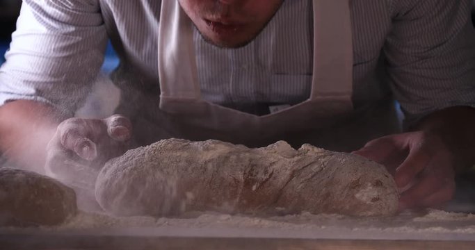 In the bakery, a baker blows off flour from bread, a lot of flour in the air. Concept of: Dirty hands, Flour, Chef, Baker.
