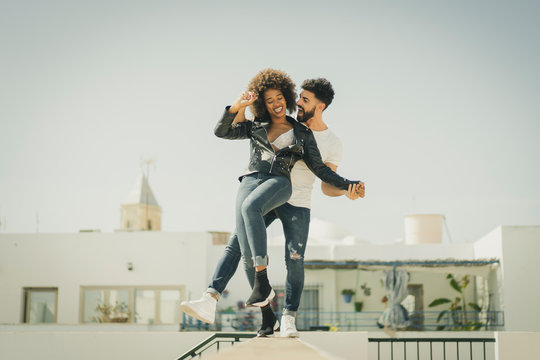 Multiracial man and woman laughing and balancing on wall while having fun on city street during date