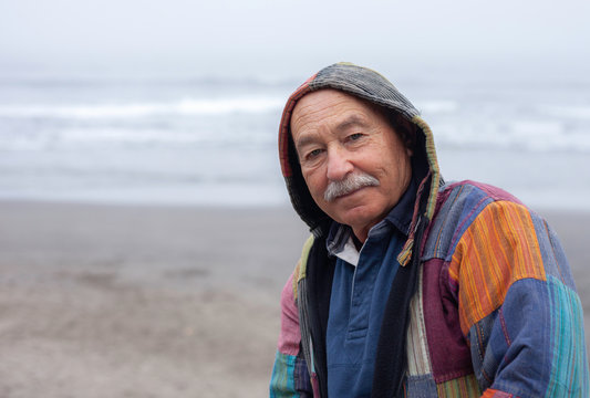 Side view of senior man in coat standing on remote empty beach of ocean coast looking at camera