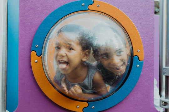 Two little African American boys looking away through porthole and making funny faces while playing on playground together
