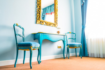 Two blue vintage chairs and a wooden table, next to a wall with a mirror, decorated with yellow floral swirls on its frame. Blue curtains on the side with soft natural light coming in from outside.