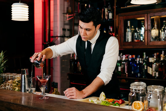 Young elegant barman working behind a bar counter pouring drink from shaker to a glass