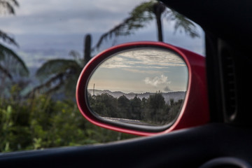 Fototapeta na wymiar View in the wing mirror of a red sports racing car. Mirror is in focus and shows clouds over a bush scene from the Waitakere Ranges in Auckland. The background is blurred tree ferns or punga.