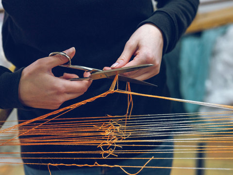 Unrecognizable weaver using scissors to cut orange cotton thread on loom on factory