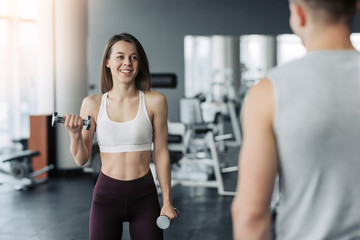 Fototapeta na wymiar Young beautiful woman doing exercises with dumbbell in gym. Glad smiling girl is enjoying with her training process. She is working hard. Her trainer looking at this process.