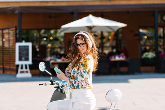 Gorgeous girl with long curly hair wearing shirt with floral pattern, sitting on scooter and holding mobile phone. Outdoor portrait of shy smiling young woman posing with moped in front of restaurant.