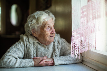 An elderly woman looks out the window sitting in his house. Care of lonely pensioners.