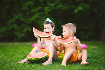 Funny little kids brother and sister eating watermelon on green grass near inflatable pool in yard at home. Toddler boy and girl. Children eat fruit in garden. Childhood, Family, Healthy Diet