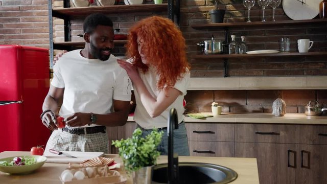 Slow motion of cheerful curly red haired woman walking to the afro American man preparing salad, she gets bottle of wine out of the shelf and offers it to her partner. Romantic atmosphere, flirting
