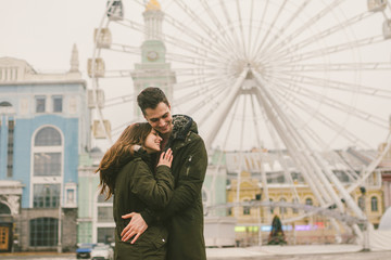 Theme love and holiday Valentines Day. pair of Caucasian heterosexual lovers in winter together gloomy weather embrace against background of Ferris wheel in town square. The guy gently hugs the girl