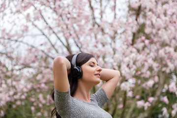 Young woman listening to music on wireless headphones in a park with cherry blossom trees.