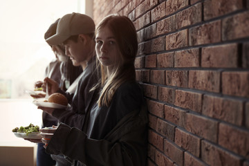 Teenage girl and other poor people with food at brick wall indoors. Space for text