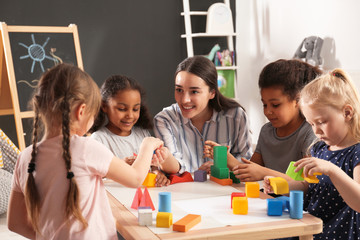 Cute little children and nursery teacher playing with building blocks in kindergarten. Indoor...