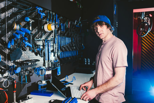 Theme business profession manager and technology. Young stylish Caucasian male owner bicycle shop uses a laptop computer near the stand with the tools for repairing and servicing the bicycle