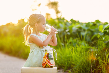 Cute little girl with blonde hair drinking lemonade outdoor. Detox fruit infused flavored water, cocktail in a beverage dispenser with fresh fruits - obrazy, fototapety, plakaty