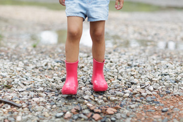 Child in a pink rubber boots in the rain jumping in puddles. Kid playing in summer park. Outdoor fun by any weather