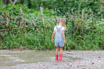 Child in a pink rubber boots in the rain jumping in puddles. Kid playing in summer park. Outdoor fun by any weather