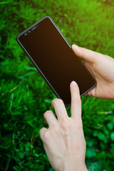 Close up young woman hand holding and touching screen on the modern black smartphone mock up in vertical position with blank screen against beautiful blurred grass background. Clipping path.