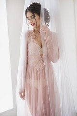 Tender young bride with wedding makeup and hairstyle in bedroom in peignoir posing in white curtains