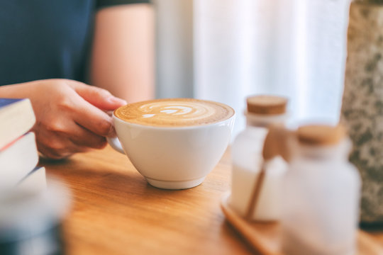 Closeup image of a woman's hand holding a white cup of hot coffee with books on wooden table