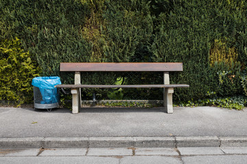 Holzbank mit angeketteter Mülltonne und blauem Müllsack vor grüner Hecke an grauer Straße