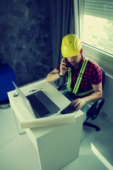 Construction Worker is sitting at his desk at the computer, looking at building plans, counting and...