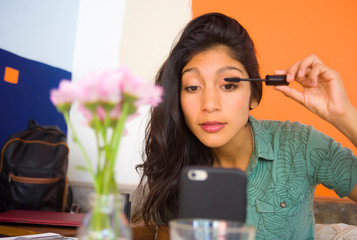 lifestyle portrait of young beautiful and attractive brunette latin woman retouching eyelashes mascara makeup using mobile phone as mirror sitting at cafe