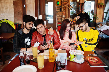 Group of asian friends sitting cafe. Happy indian people having fun together, sitting on couch and looking at mobile phones.