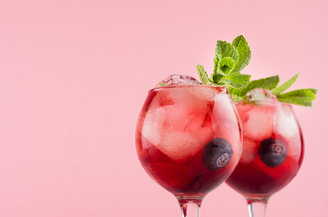 Fresh cold fruit cocktails in two elegant goblets with ice cubes, blueberry, green mint closeup, top section on pastel pink color background, copy space.