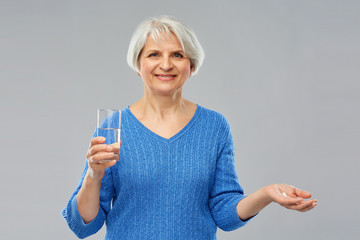 medicine, health and old people concept - smiling senior woman with glass of water and pills over grey background