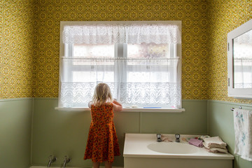 Young girl alone exploring the bathroom of a 1970s house. Lace curtains and mustard yellow colored...