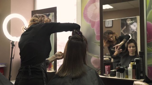 young curly stylist in apron trims and straightens beautiful dark hair ends with silver scissors and comb close view