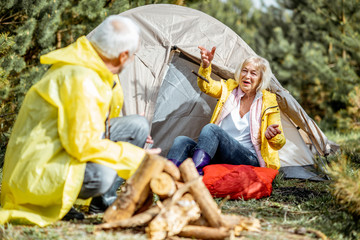 Senior couple in yellow raincoats having fun while making fireplace at the campsite near the tent in the woods