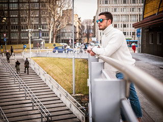 One attractive man in urban setting in modern city, leaning on metal handrail, wearing wool sweater and sunglasses, looking away