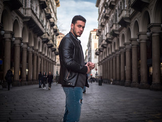 Fototapeta na wymiar One handsome young man in urban setting in old classic European city, standing, wearing black leather jacket and jeans, looking at camera in Turin, Italy