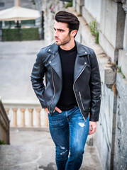 Obraz premium One handsome young man in urban setting in modern city, standing, wearing black leather jacket and jeans, looking away